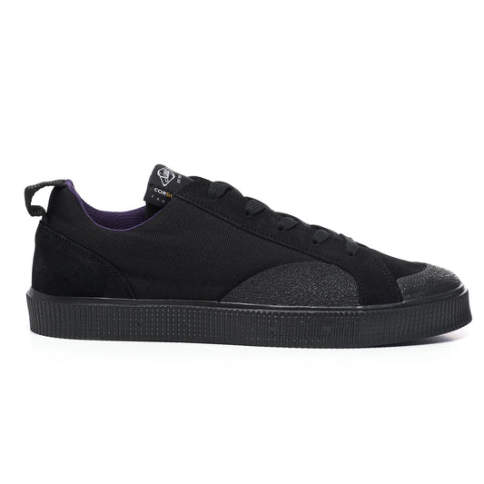 A side view of a Sanjo TESTE low-top sneaker featuring a sleek design with a black Cordura nylon upper for durability and water resistance. Accented with suede overlays on the heel and toe for added texture. Includes a pull tab on the heel for easy wear and a tonal rubber outsole with textured detailing for grip. Finished with a subtle logo on the tongue and a contrasting purple inner lining.